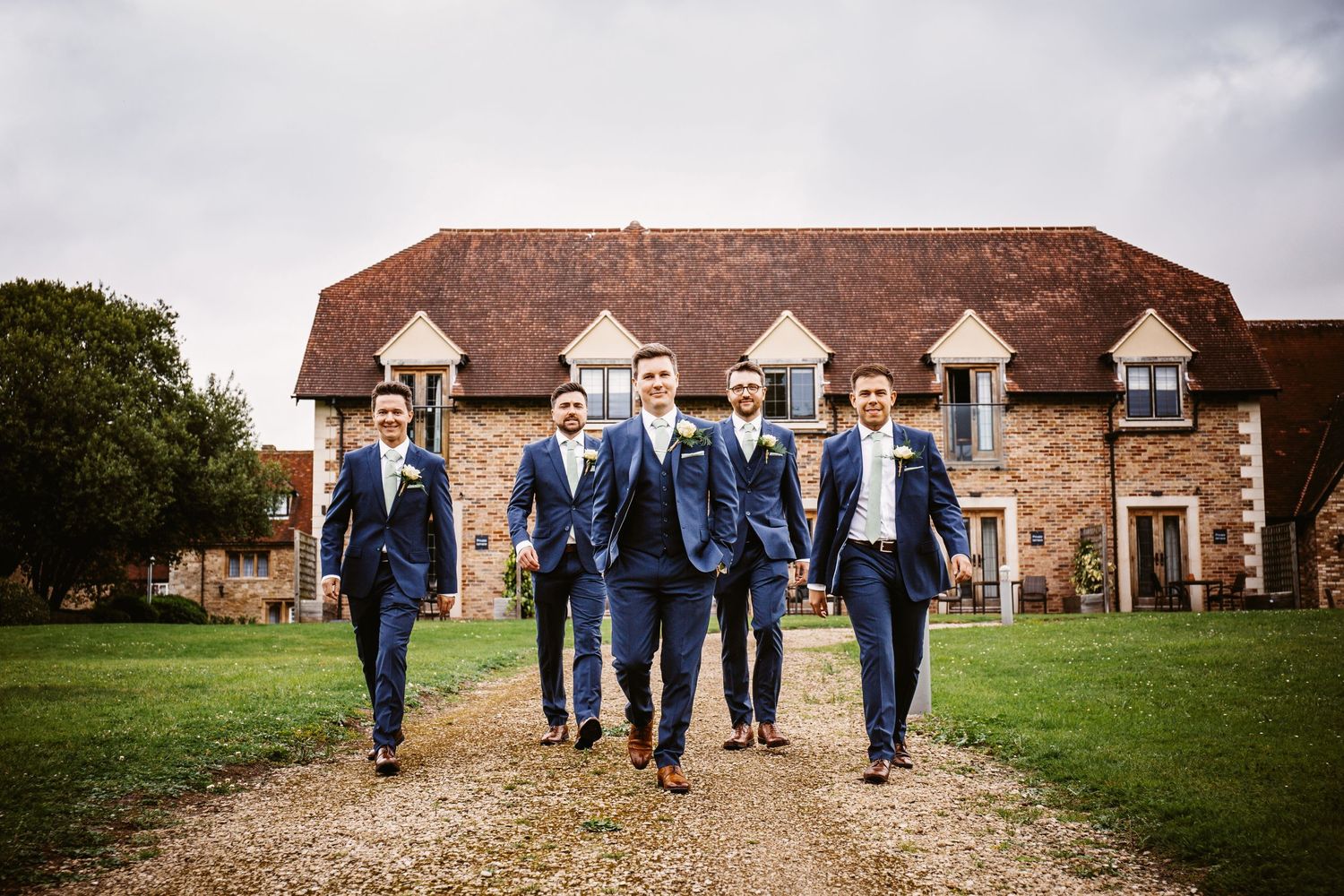 Wedding party in navy suits walking along a gravel path towards a rustic manor house with dormers.