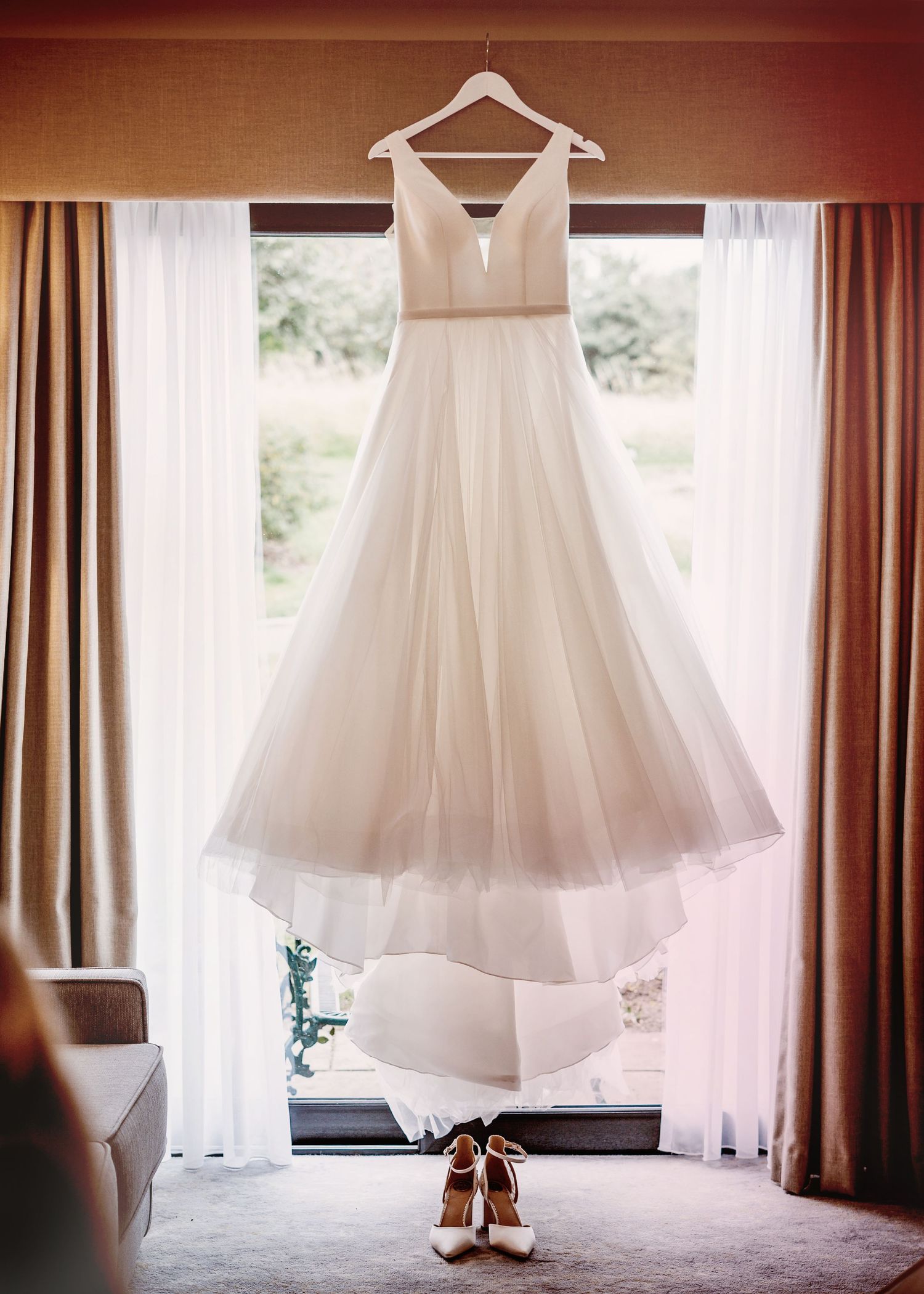 Elegant white wedding gown hanging in window with tulle skirt and bridal shoes below.