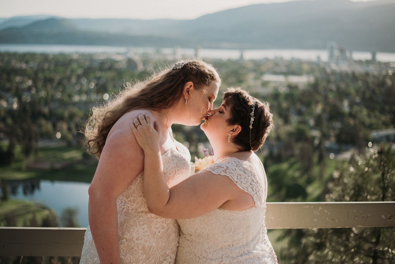 Wedding photo of brides kissing with the view of Kelowna behind.