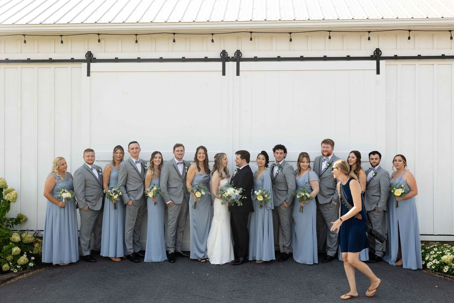 Large wedding party in gray and blue attire posed in front of white barn wall.