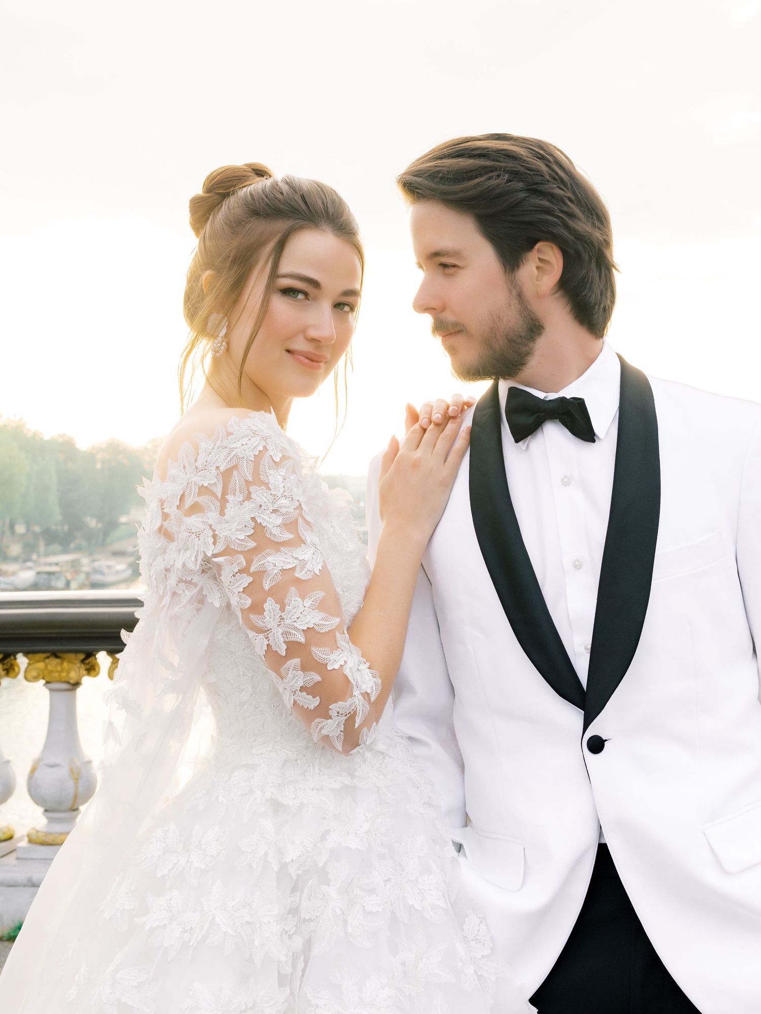 An elegant wedding portrait featuring a white tuxedo and lace dress against a sunset backdrop.