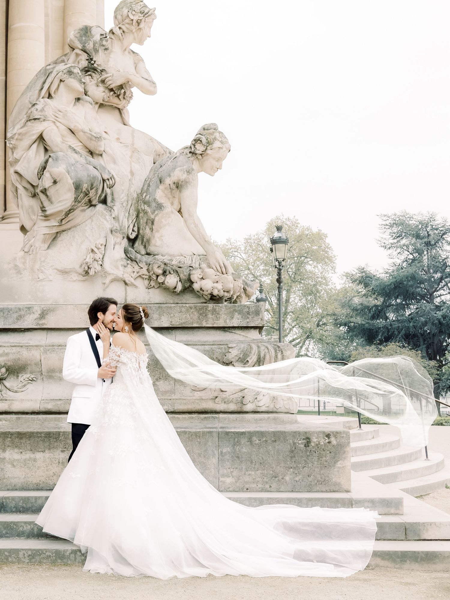 A romantic wedding couple embracing near ornate stone statues and architecture with a flowing white veil and dress.