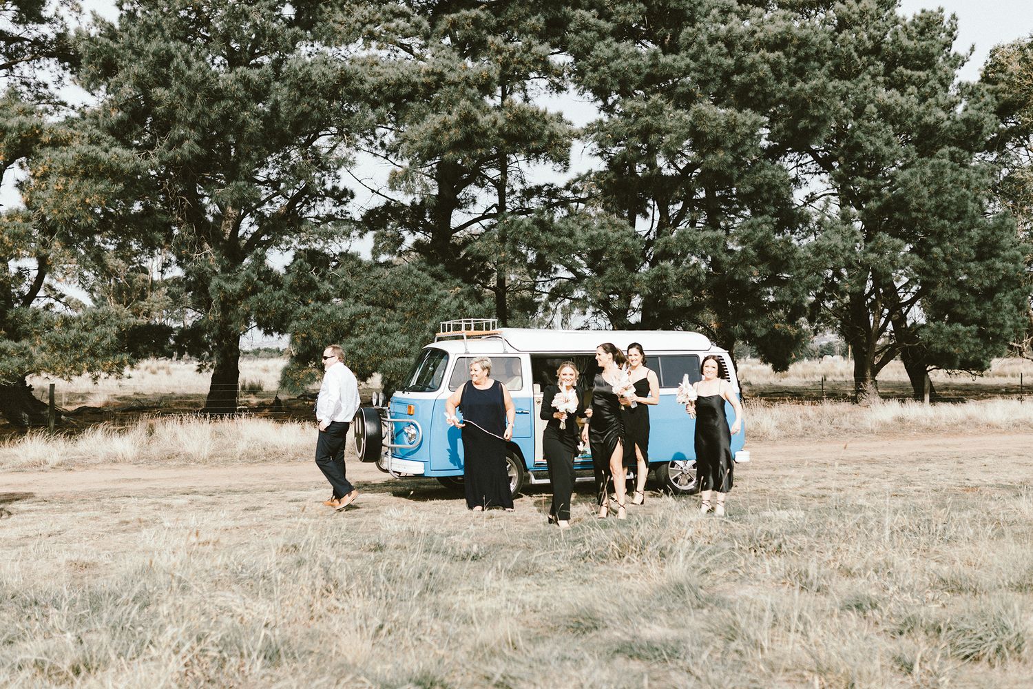 Wedding group gathers around vintage blue and white van in rustic setting.