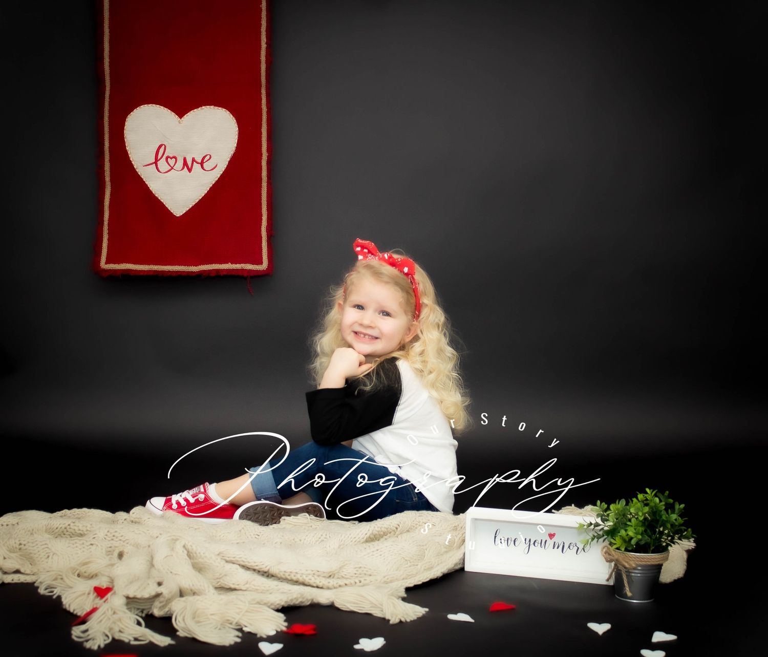 A young child in white sits against a black backdrop with a red love banner and Valentine's Day decor.