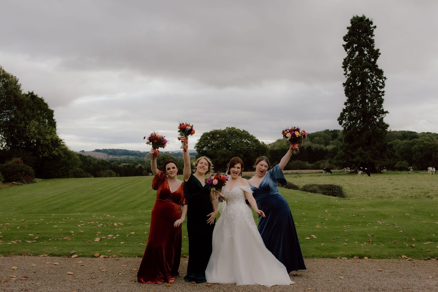 Group celebrates on grassy lawn with raised bouquets against cloudy sky.
