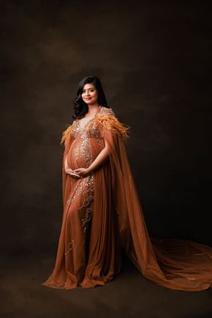 Expectant mother poses in glamorous copper-colored gown with sheer cape and feather details against dark backdrop.