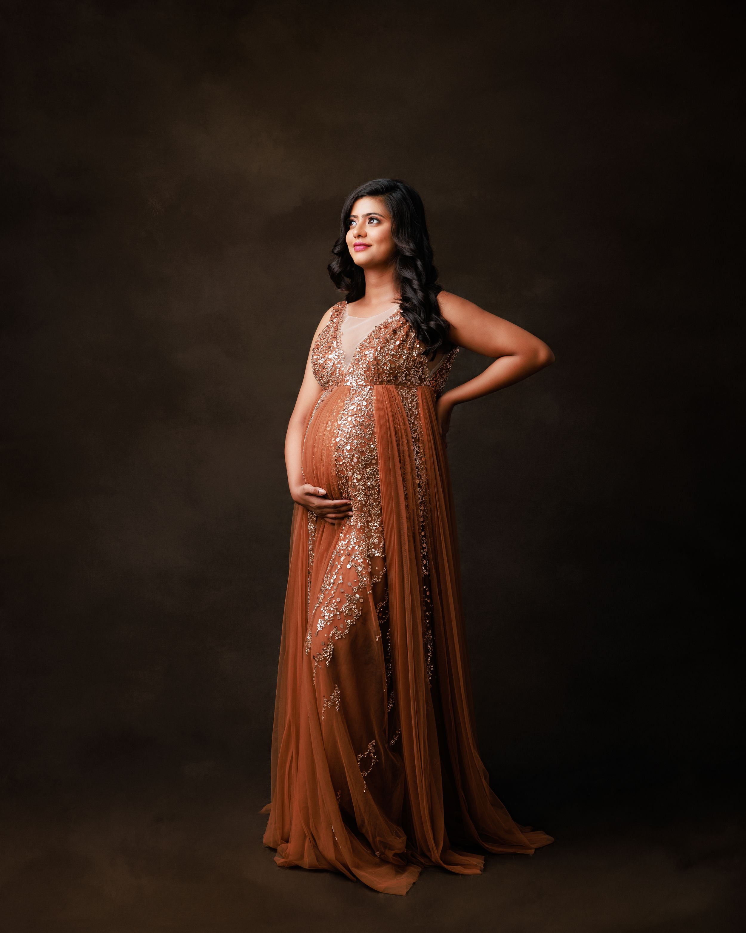 Elegant maternity portrait in a copper-toned embellished gown against dark background.