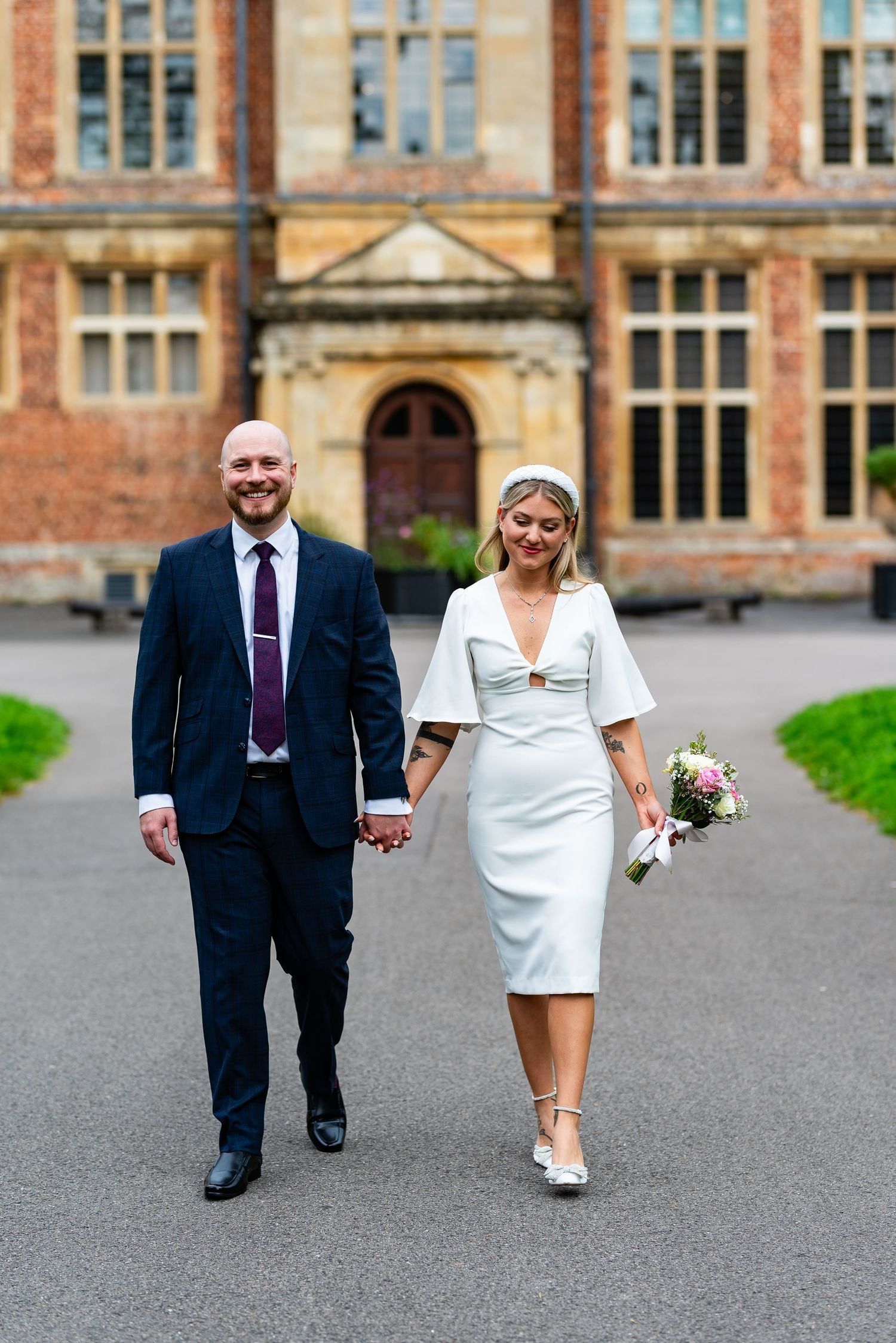 A couple wearing wedding attire walks hand in hand in front of an elegant brick building.