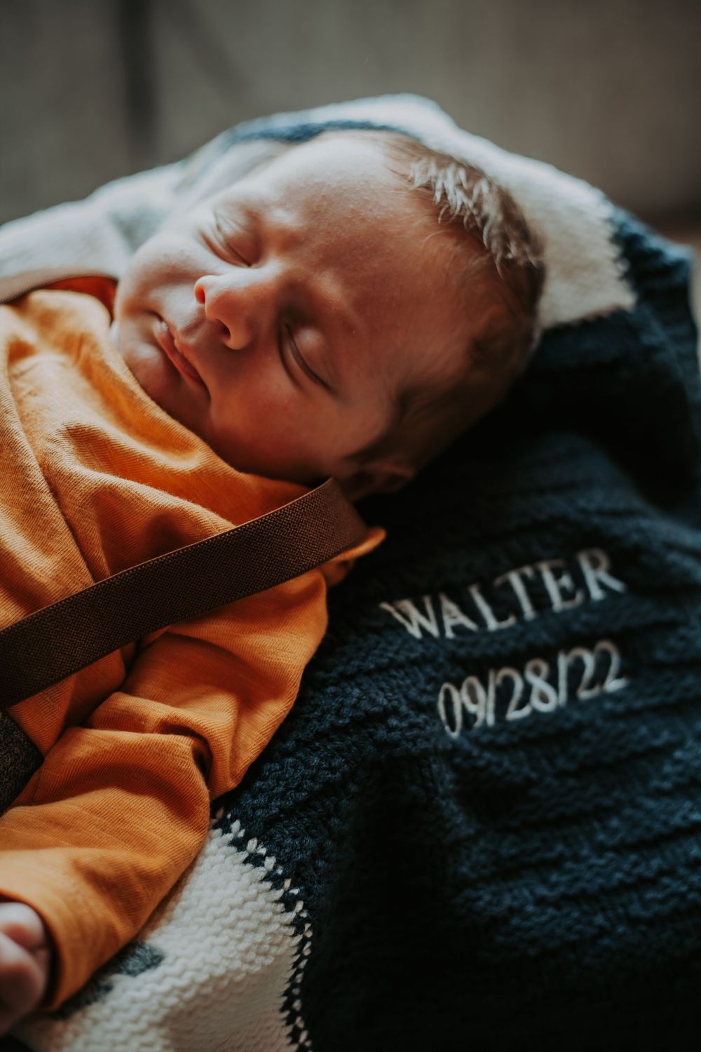 Sleeping newborn in an orange outfit rests on a navy knit blanket embroidered with the name Walter and date 09/28/22.