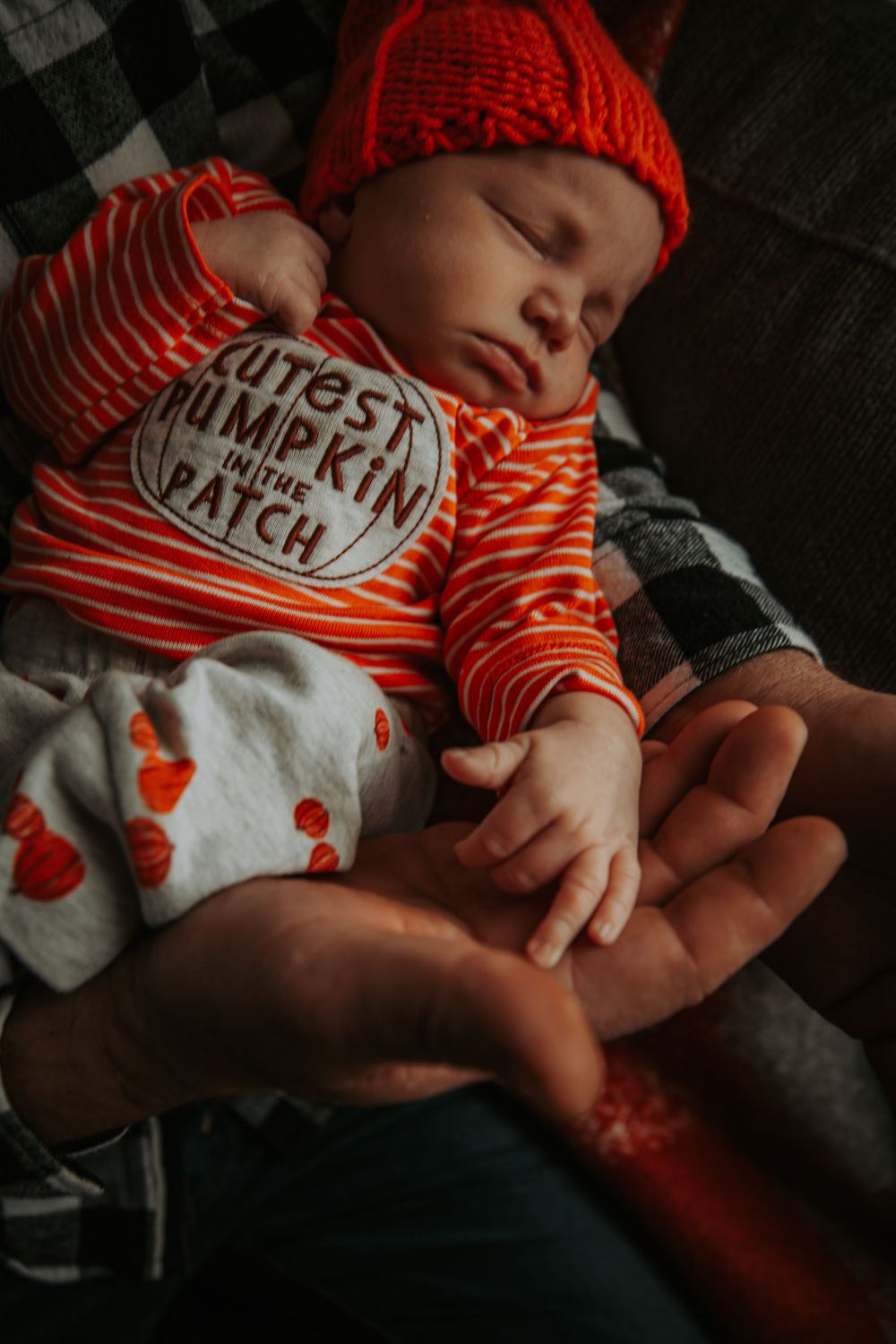 Sleeping newborn in an orange knit hat and Cutest Pumpkin in the Patch outfit rests in a father's hands.