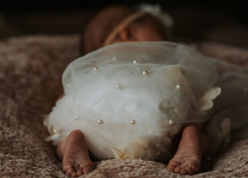 Newborn baby in a white tulle dress with pearl details lies on a soft textured blanket with tiny feet in the foreground.