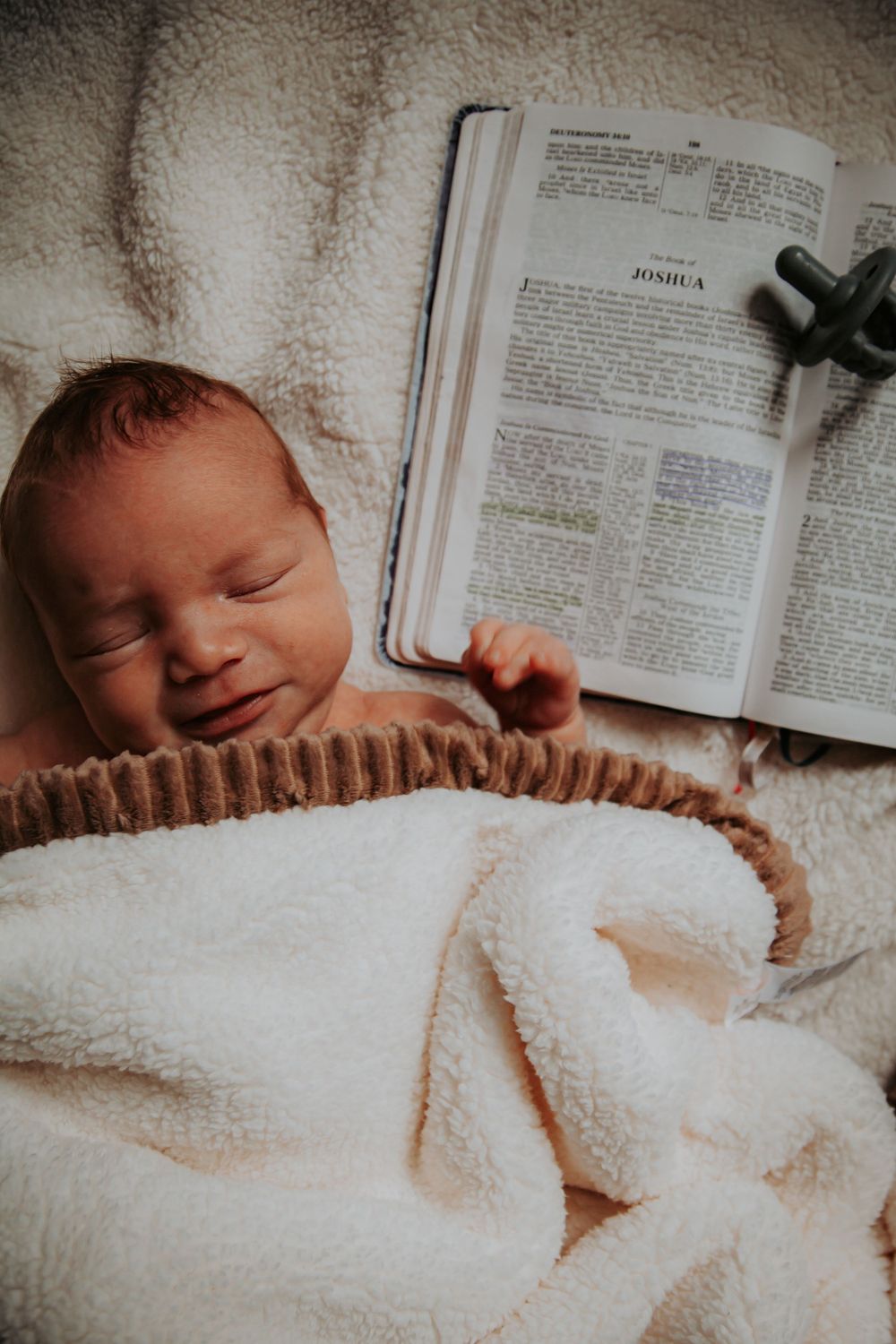 Sleeping newborn swaddled in a soft white sherpa blanket beside an open Bible and a pacifier.