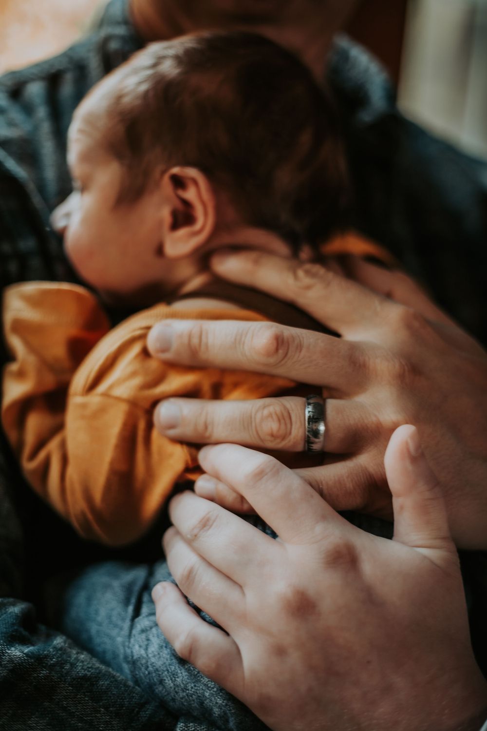 Parent's hands with a wedding ring gently cradle a sleeping newborn baby dressed in a mustard yellow top.