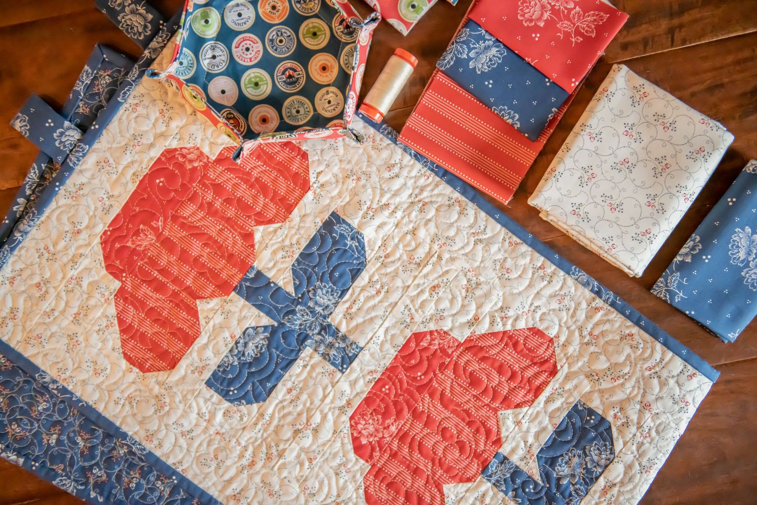 Quilted wall hanging featuring red flowers with blue stems on cream background.
