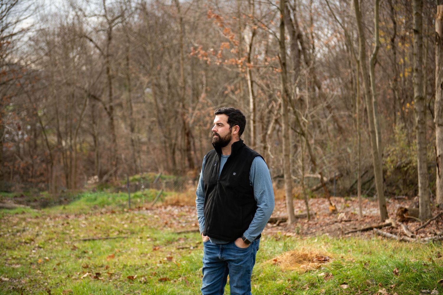 A person wearing a black vest and blue shirt stands in a wooded area during autumn.