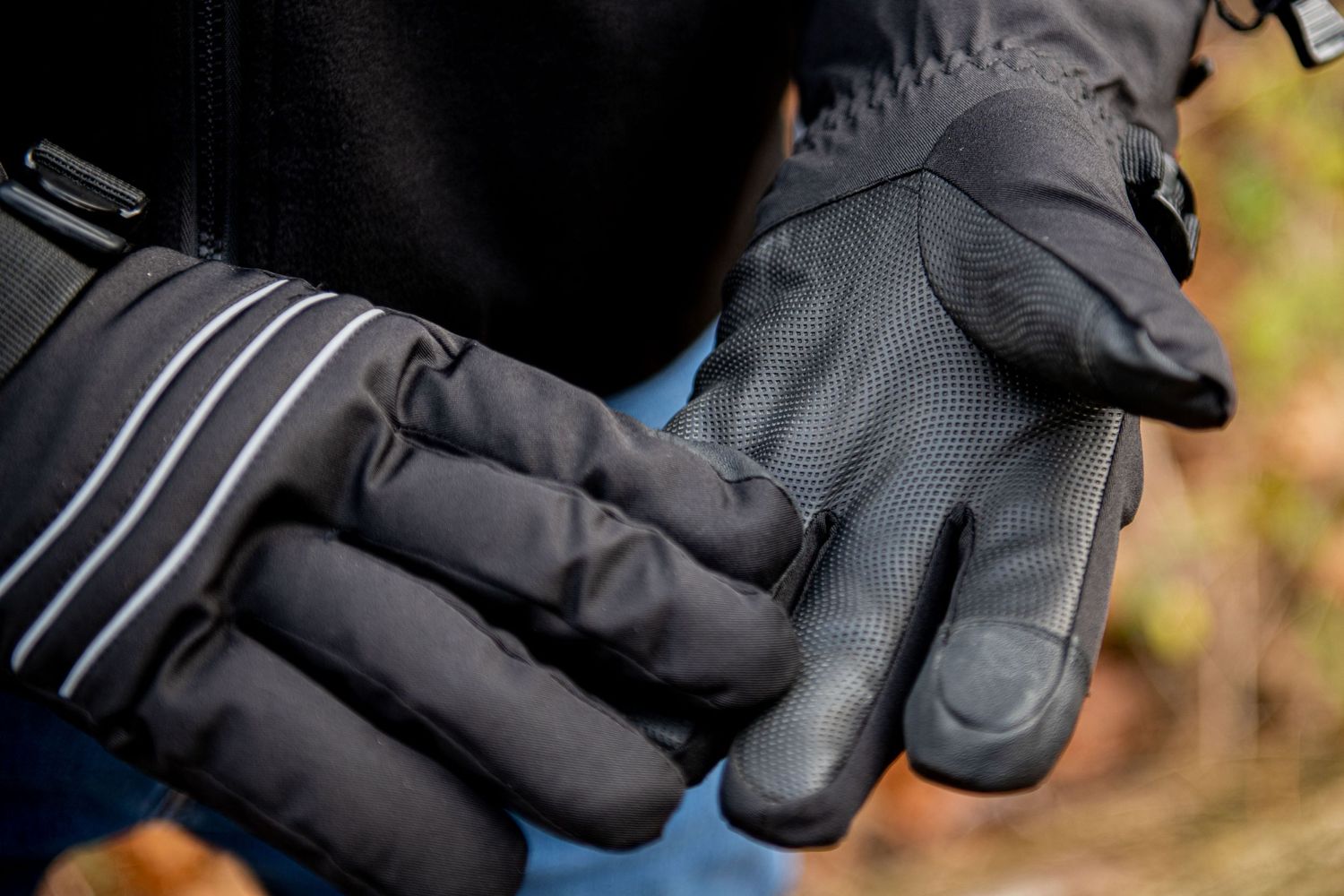 Close-up of black winter gloves with reflective stripes against a blurred outdoor background.