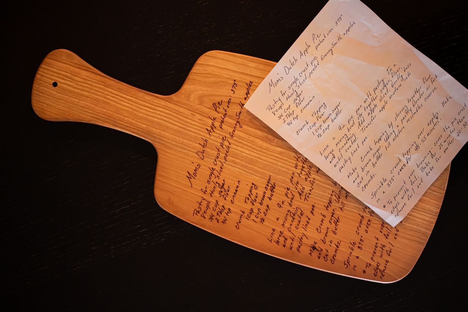 Wooden cutting board with handwritten recipe text engraved on its surface against dark background.
