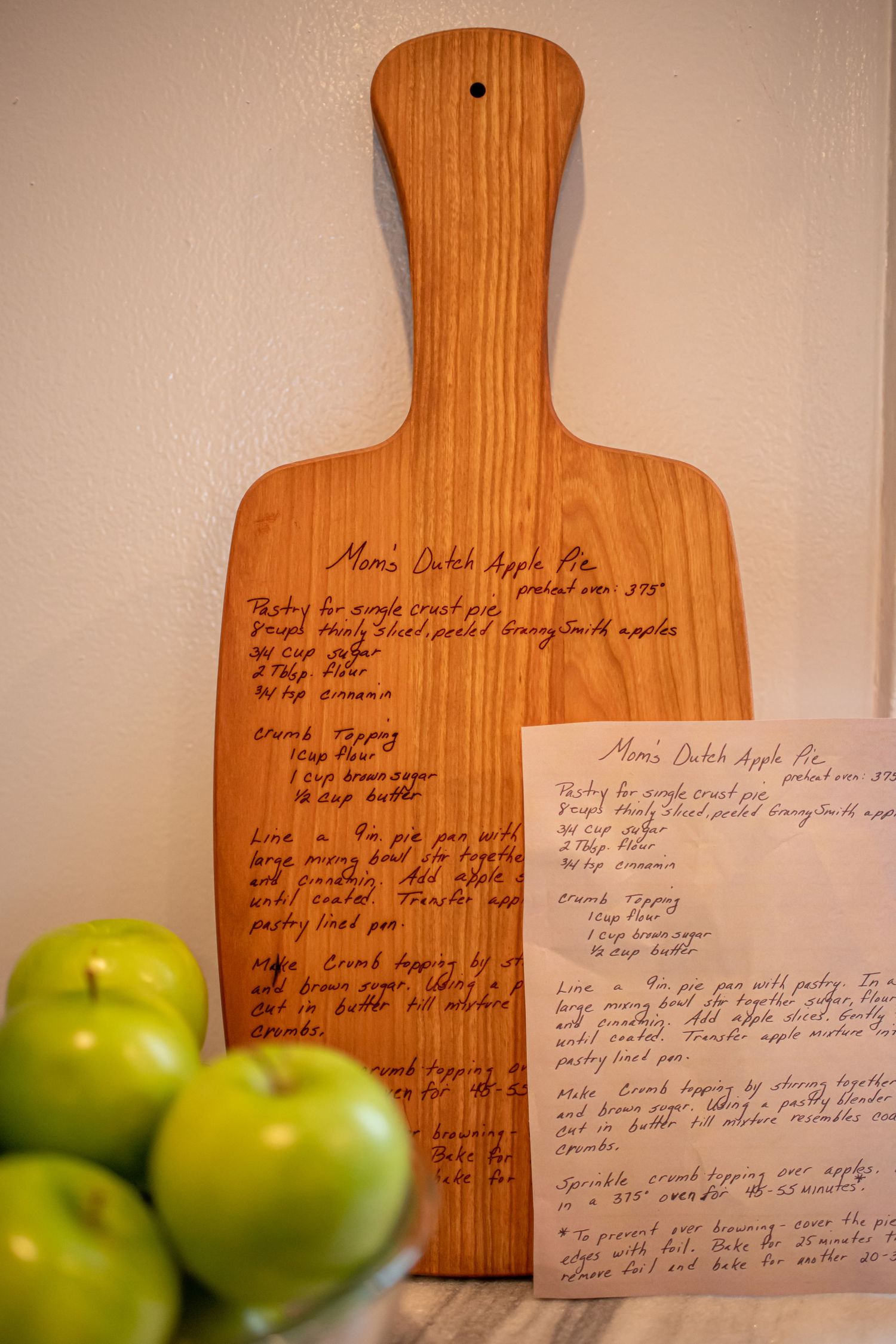 Wooden cutting board with handwritten recipe next to green apples.