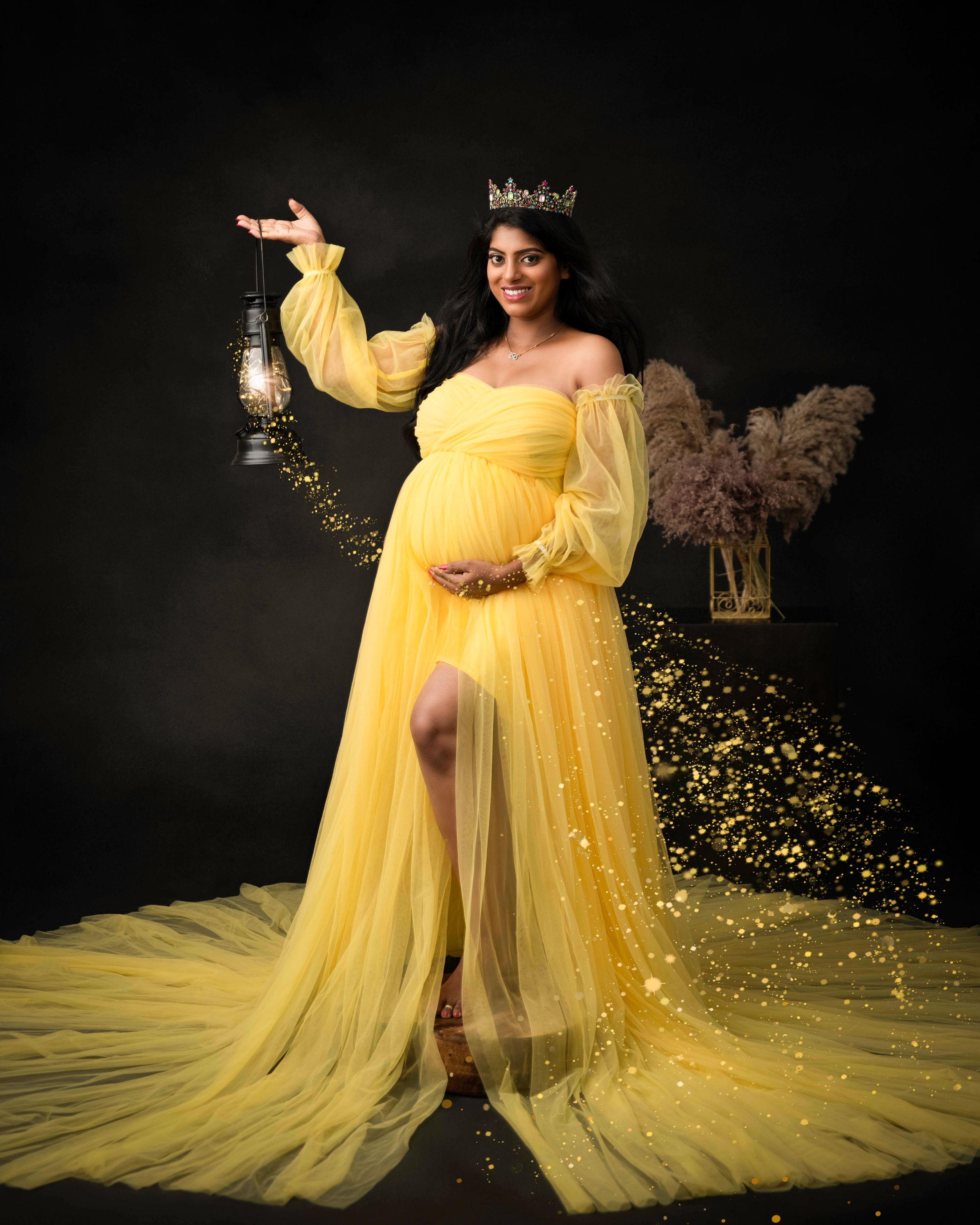 Ethereal maternity portrait in yellow gown holding lantern with floating glitter effects.