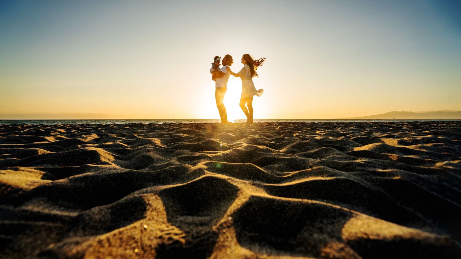 Mother, father and baby silhouettes dancing on textured sand with the sun setting behind them.