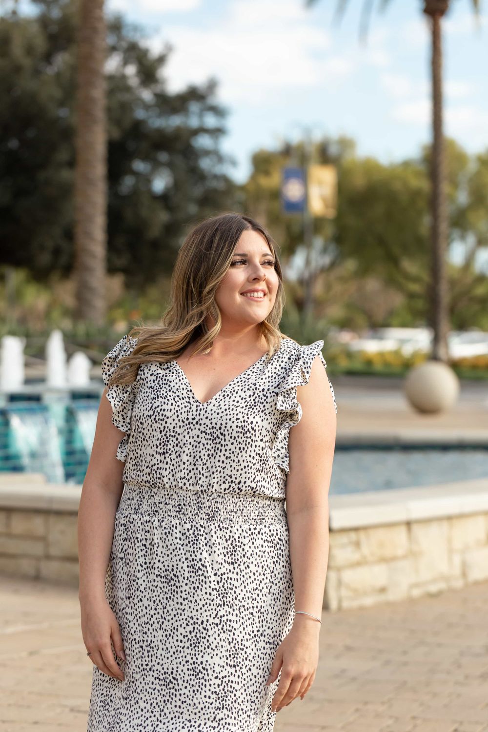 Smiling woman in white dalmatian print dress standing near fountain with palm trees in background.