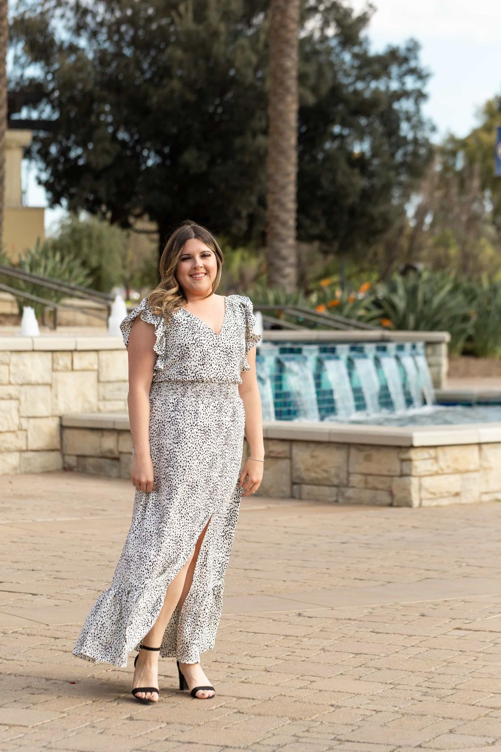 Woman in white floral maxi dress with ruffle sleeves poses near fountain outdoors.