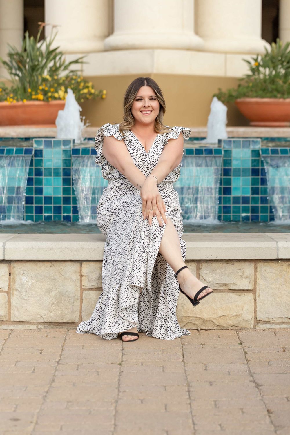 Smiling woman in spotted maxi dress sits by blue tiled fountain outdoors.
