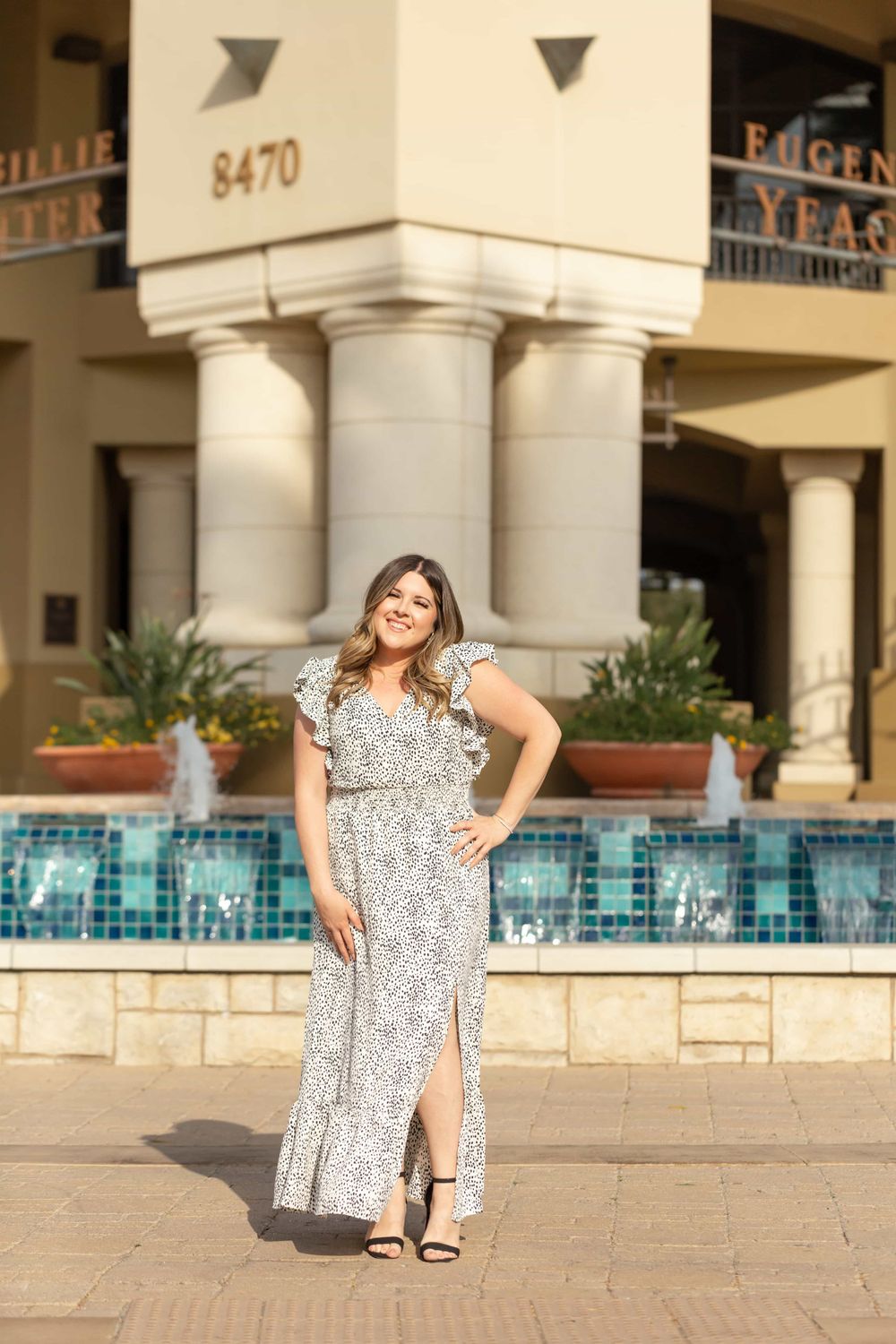 Woman in white dalmatian print maxi dress poses confidently by outdoor fountain.