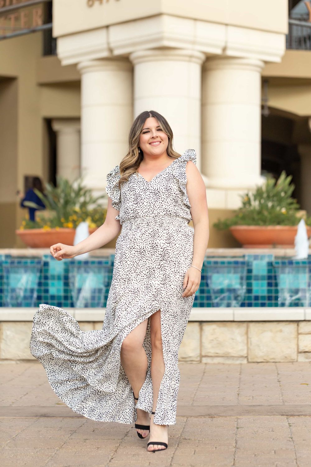 Woman twirling white spotted maxi dress with ruffle sleeves near blue tiled fountain.