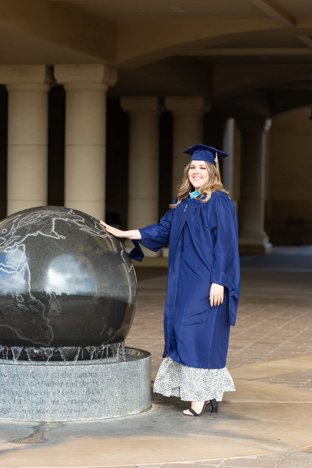 Graduate in navy cap and gown poses beside large globe sculpture in columned hall.