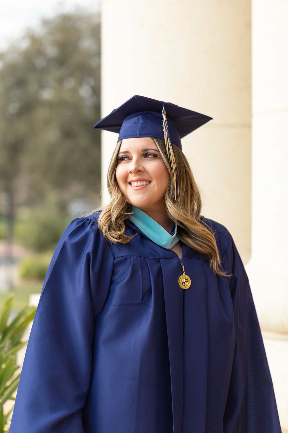 Smiling graduate in navy cap and gown with teal hood stands between white columns.