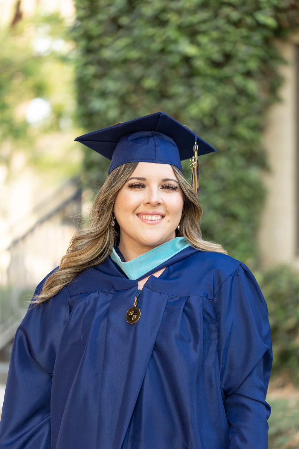 Smiling woman in navy blue graduation cap and gown with teal hood outdoors.