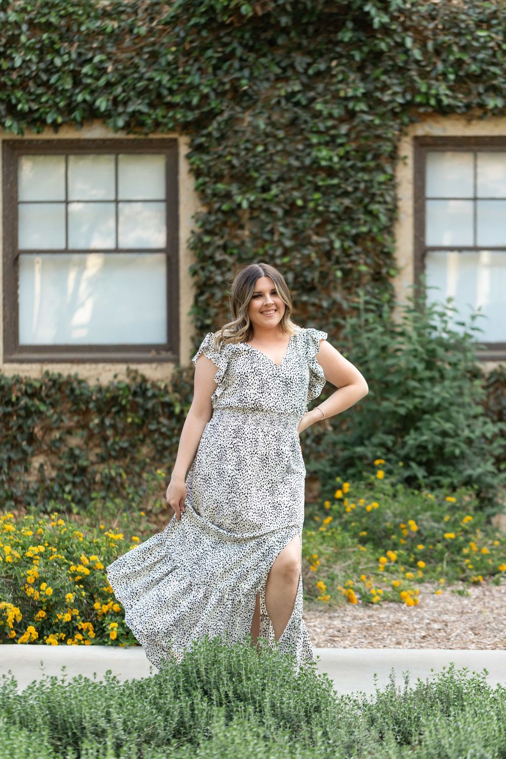 Woman in white dalmatian print maxi dress posing before ivy-covered stone building.