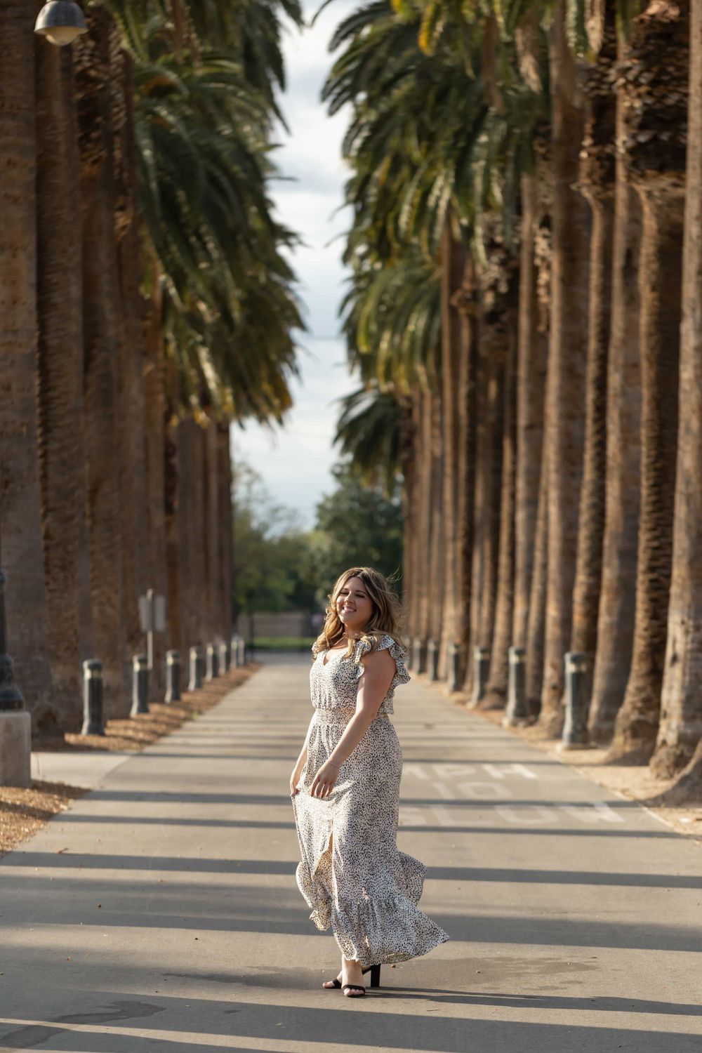 Woman in floral maxi dress walking along palm tree-lined pathway in sunlight.