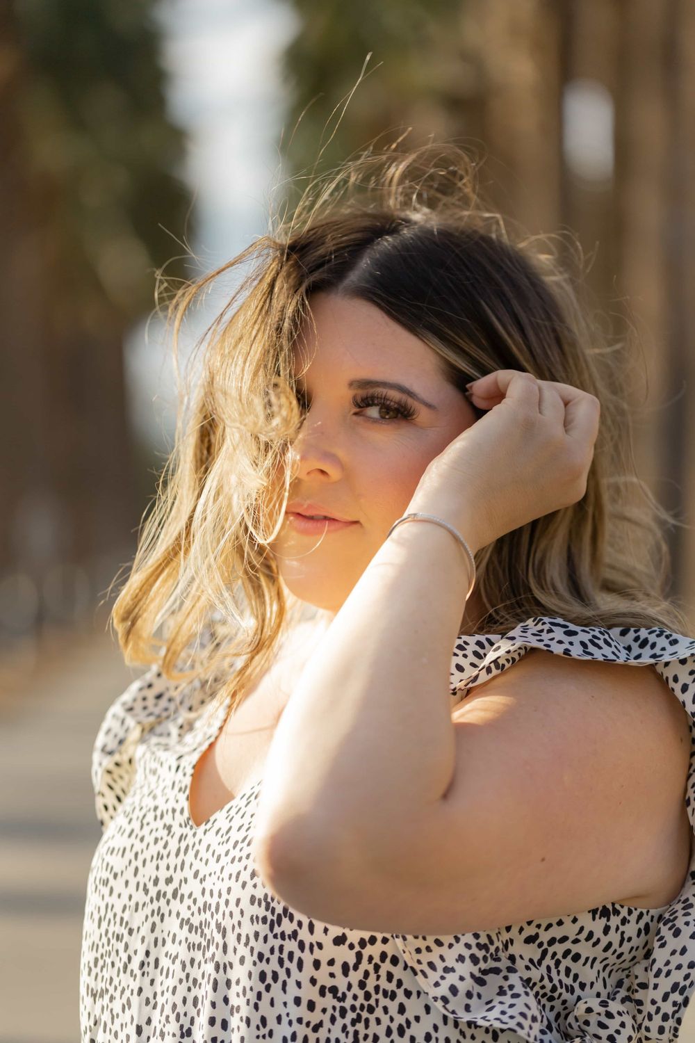 Close-up portrait of blonde woman in spotted dress with sunlit bokeh background.