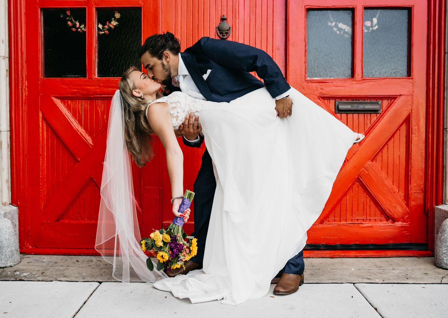 A groom in a dark suit boldly dips and kisses his bride with her colorful bouquet and white veil flowing in the wind