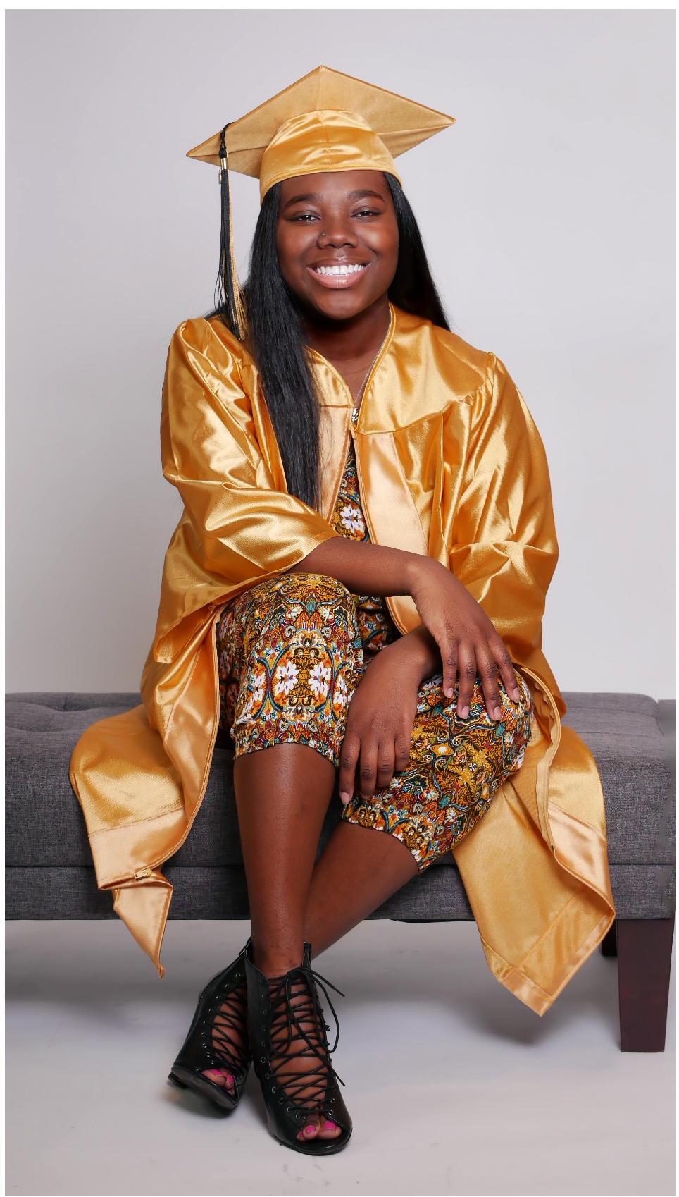 Graduate poses in gold cap and gown with floral dress on bench.