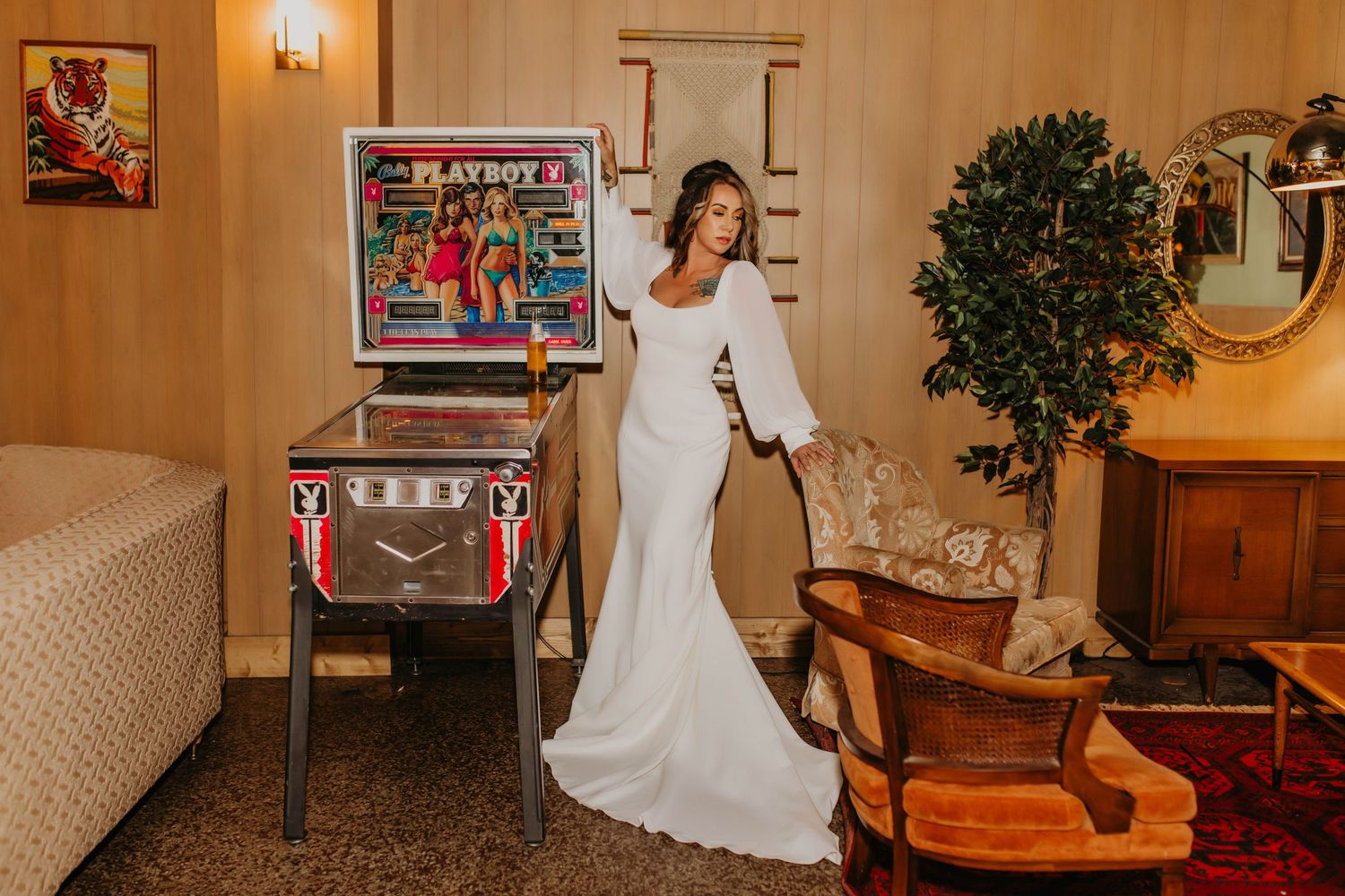 Bride in white long-sleeved gown poses next to vintage pinball machine in retro-styled room.