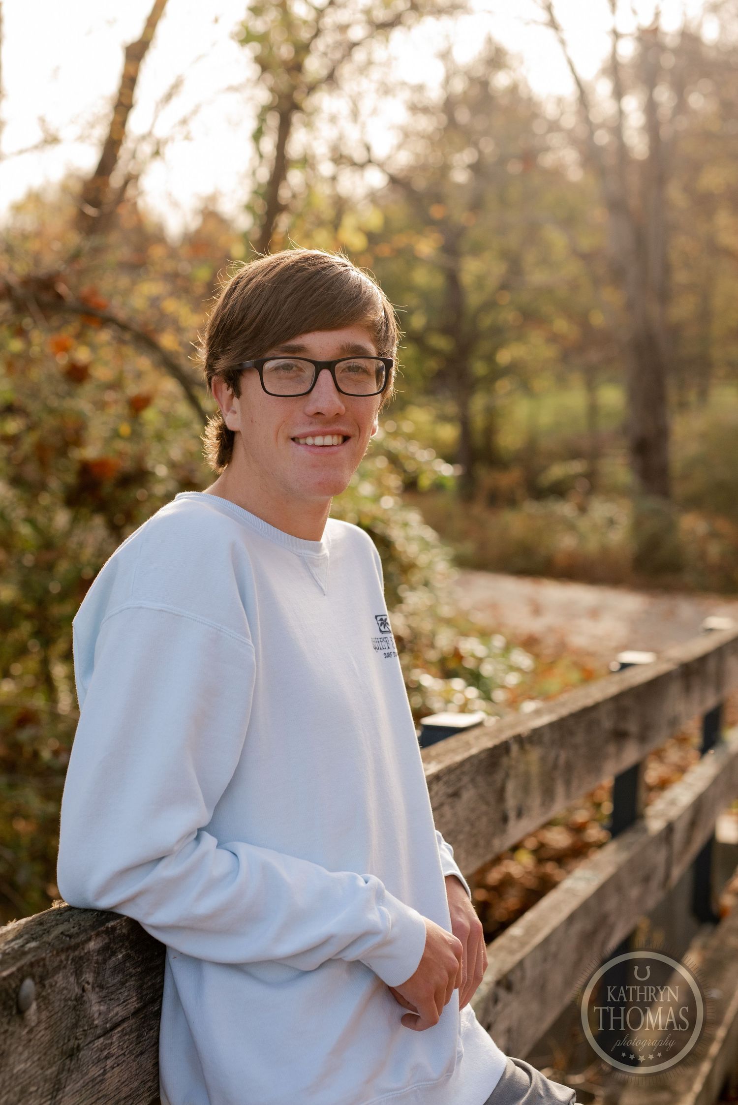 Student poses on rustic bridge railing during golden hour in autumn setting.
