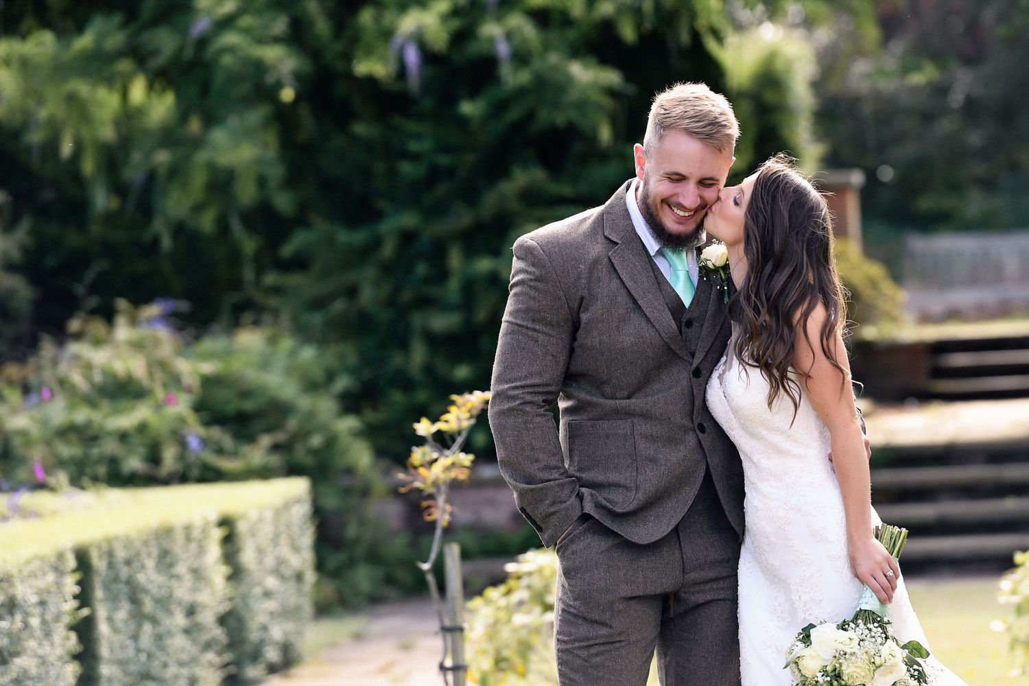 Newlyweds share an intimate moment in a garden setting with stone walls and greenery.
