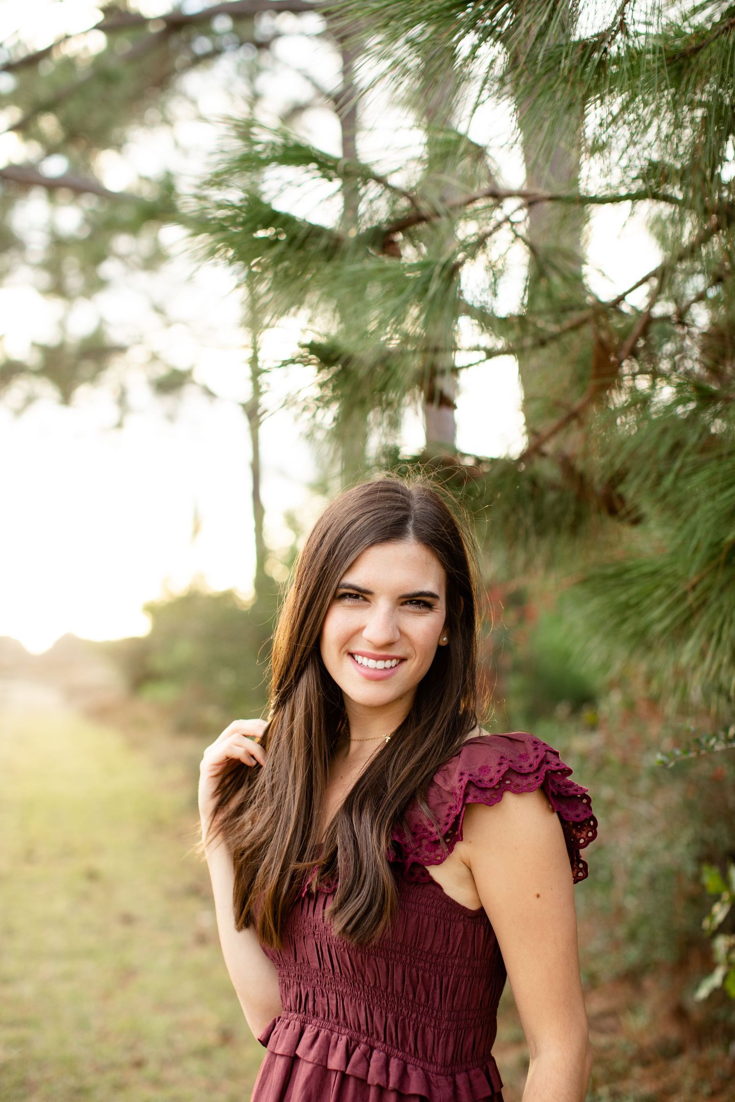 Woman wearing burgundy ruffle dress poses under pine branches at sunset.