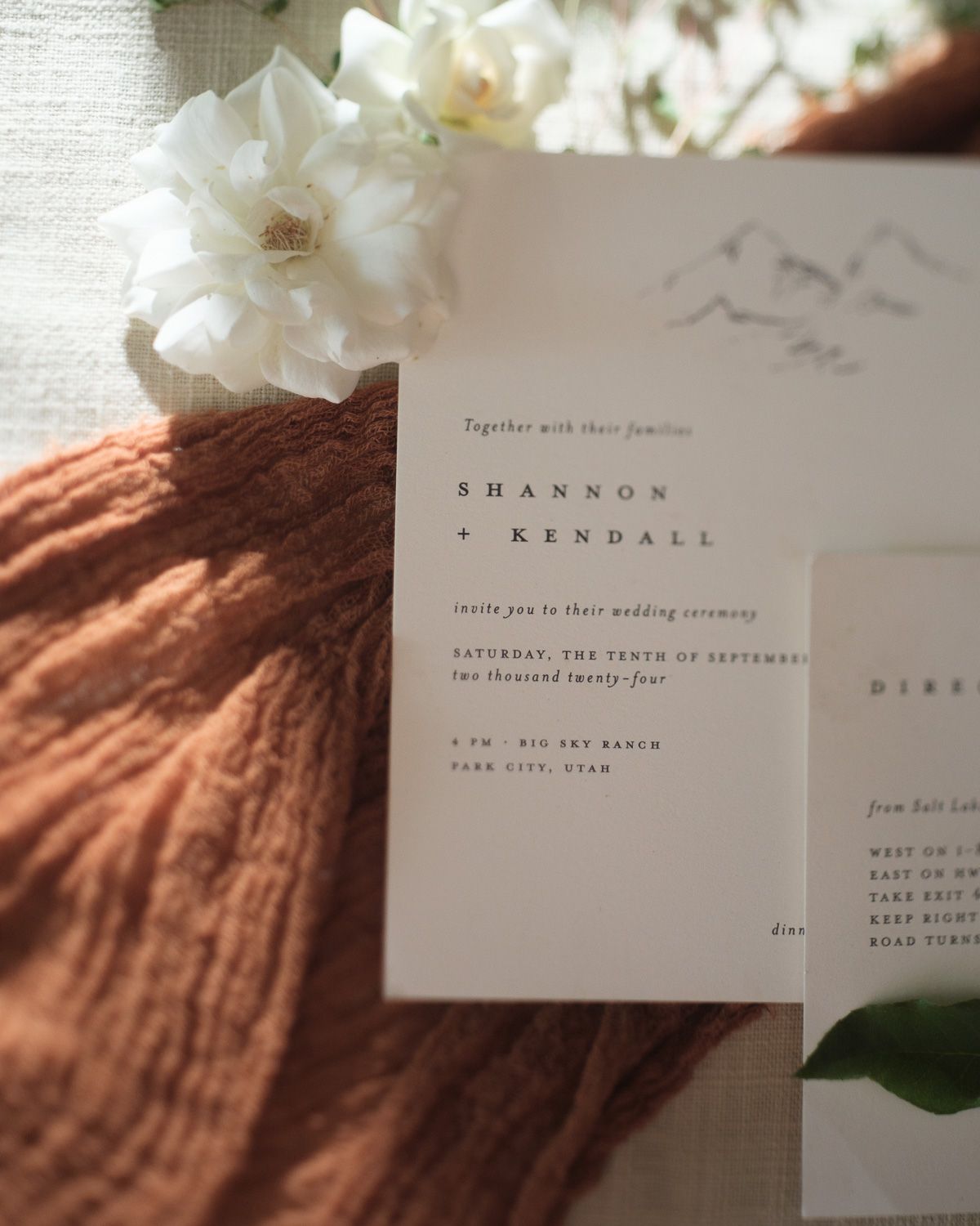 A minimalist wedding invitation with flowy text rests against an orange linen fabric, adorned with white flowers.