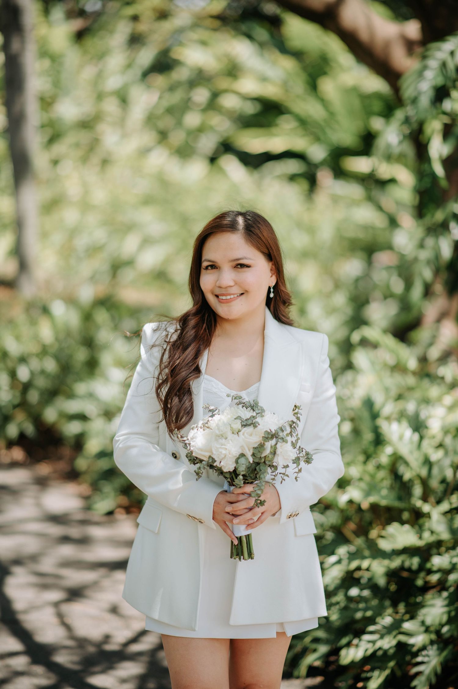 A bride in a chic white blazer dress holds a delicate bouquet of white flowers in a garden setting.