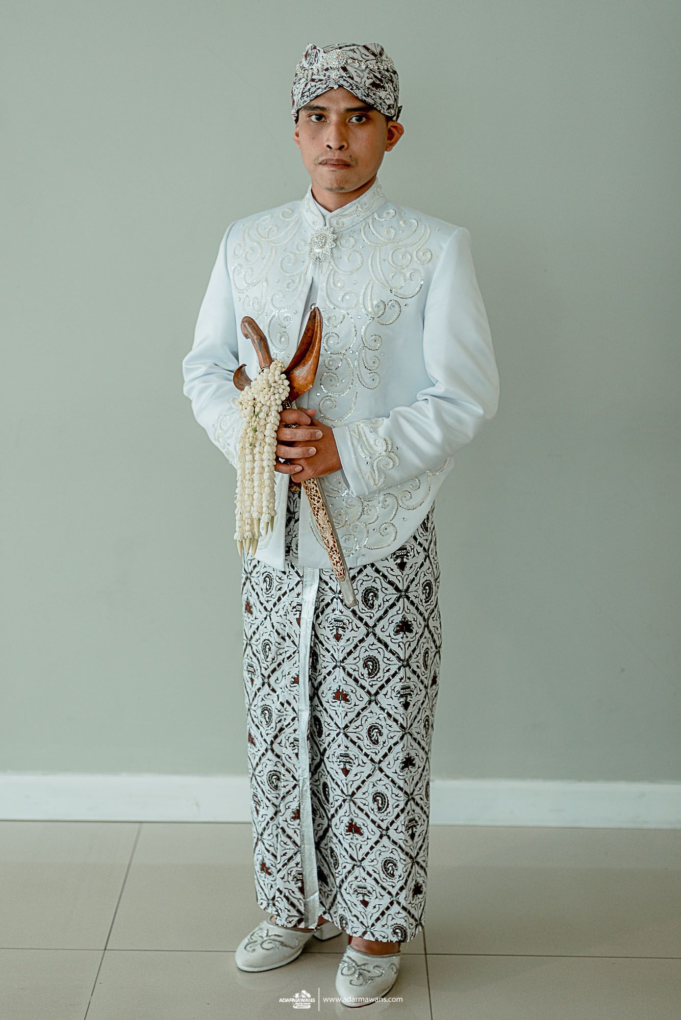 Groom in traditional Indonesian white wedding attire with decorative pattern and headdress.