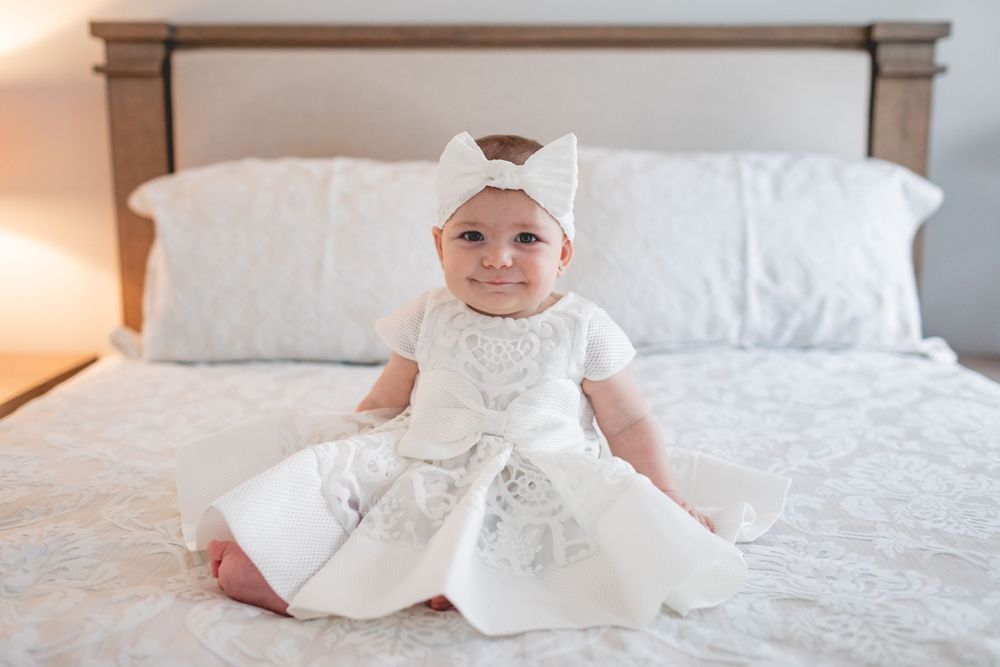 A baby in an ornate white christening gown and bonnet sits on a bed.