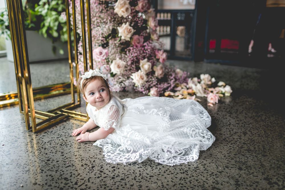 A delicate lace christening gown spreads across the floor near floral arrangements and gold accents.