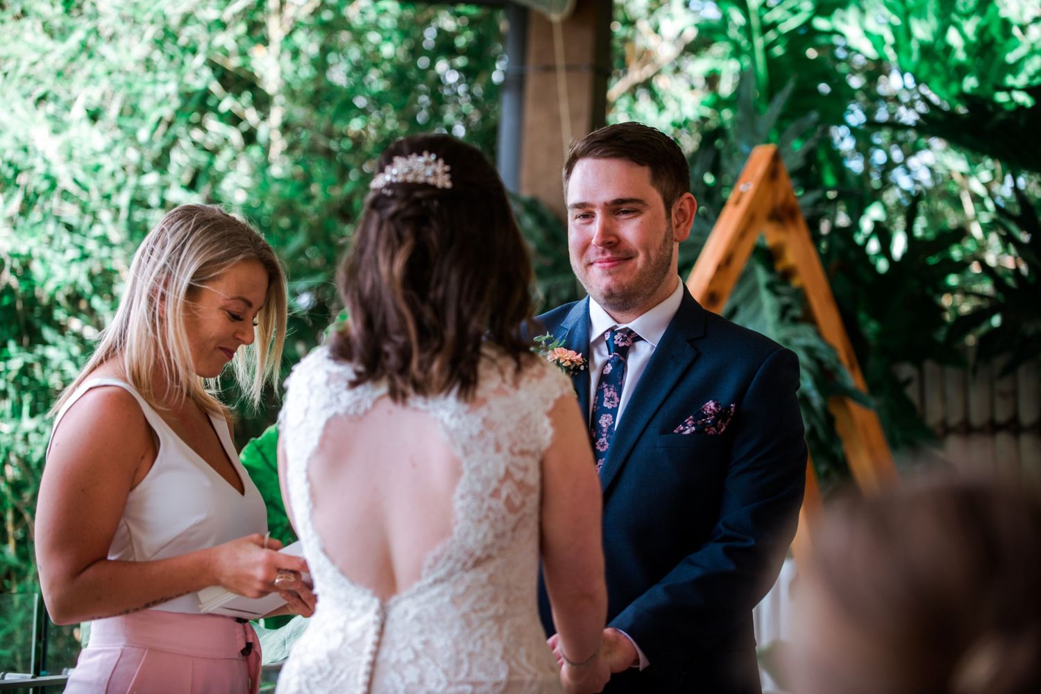 Wedding ceremony scene under wooden A-frame with greenery backdrop.