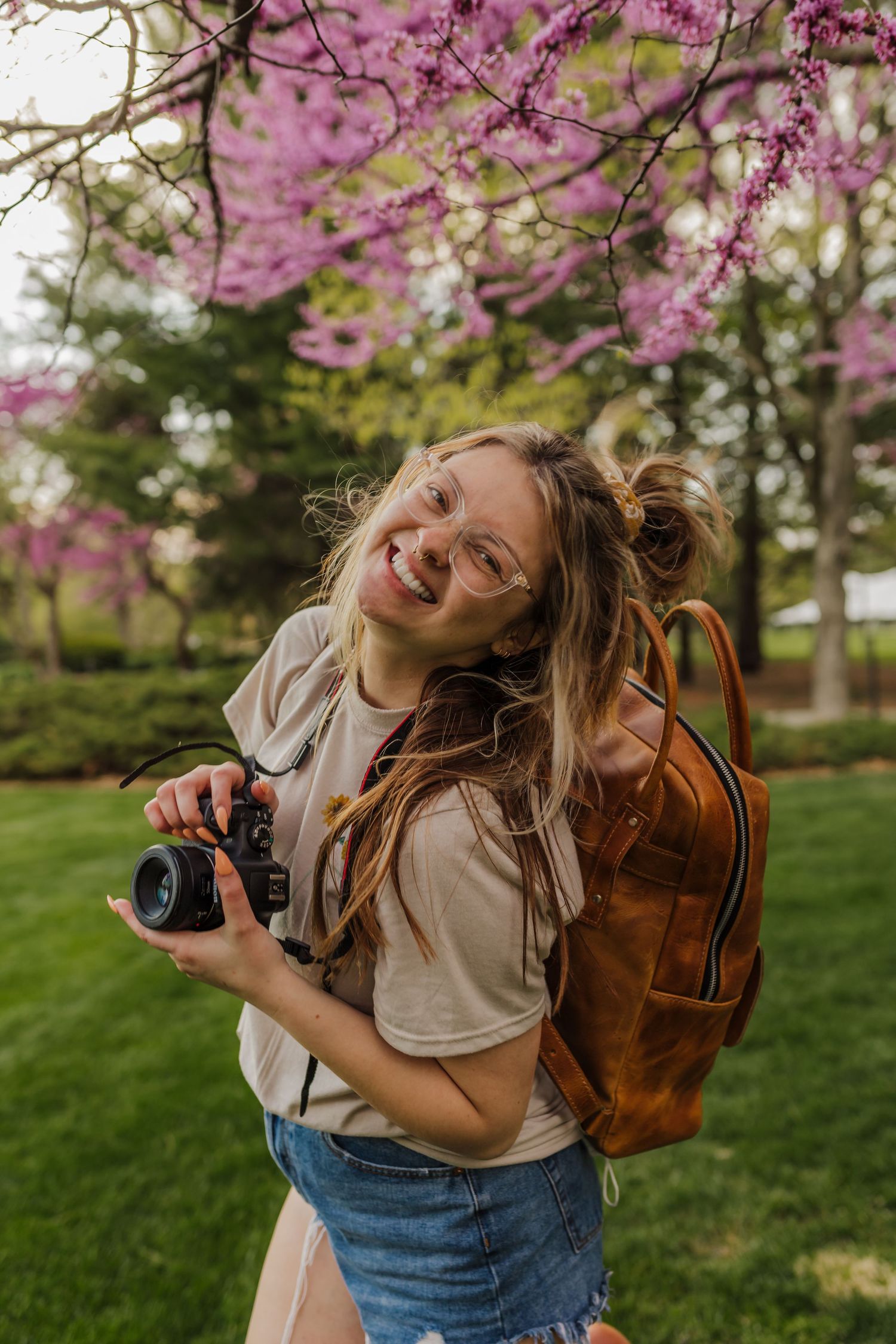 A joyful photographer with a camera and brown backpack poses under blooming cherry blossoms in a spring garden.