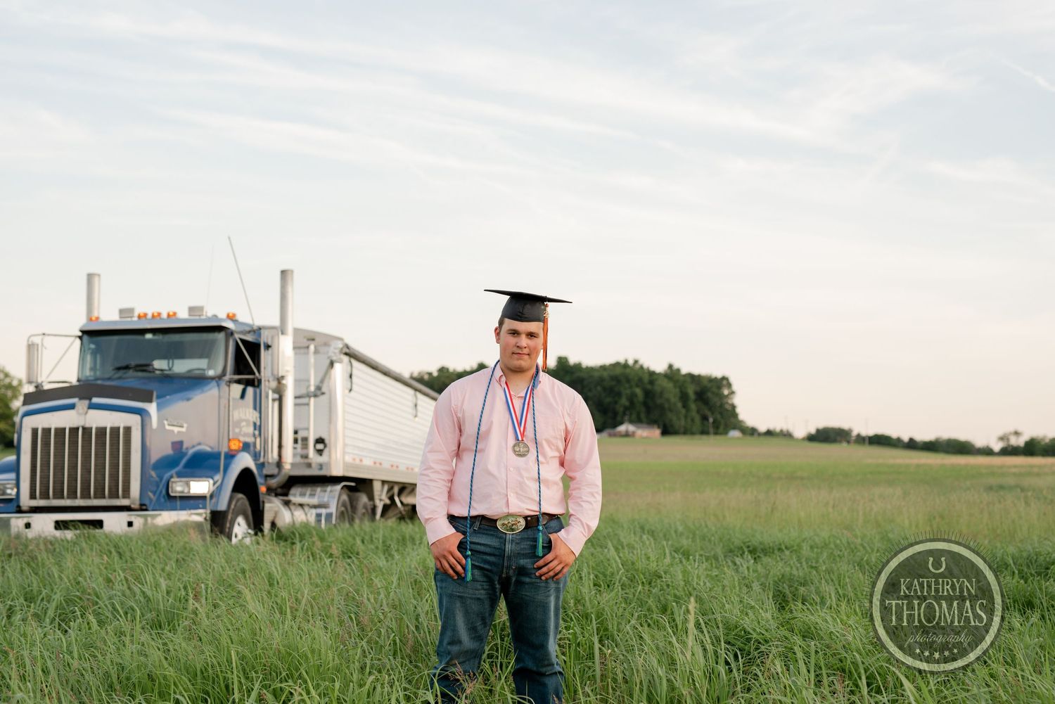 Graduate in cap and gown stands in grassy field with semi truck behind.