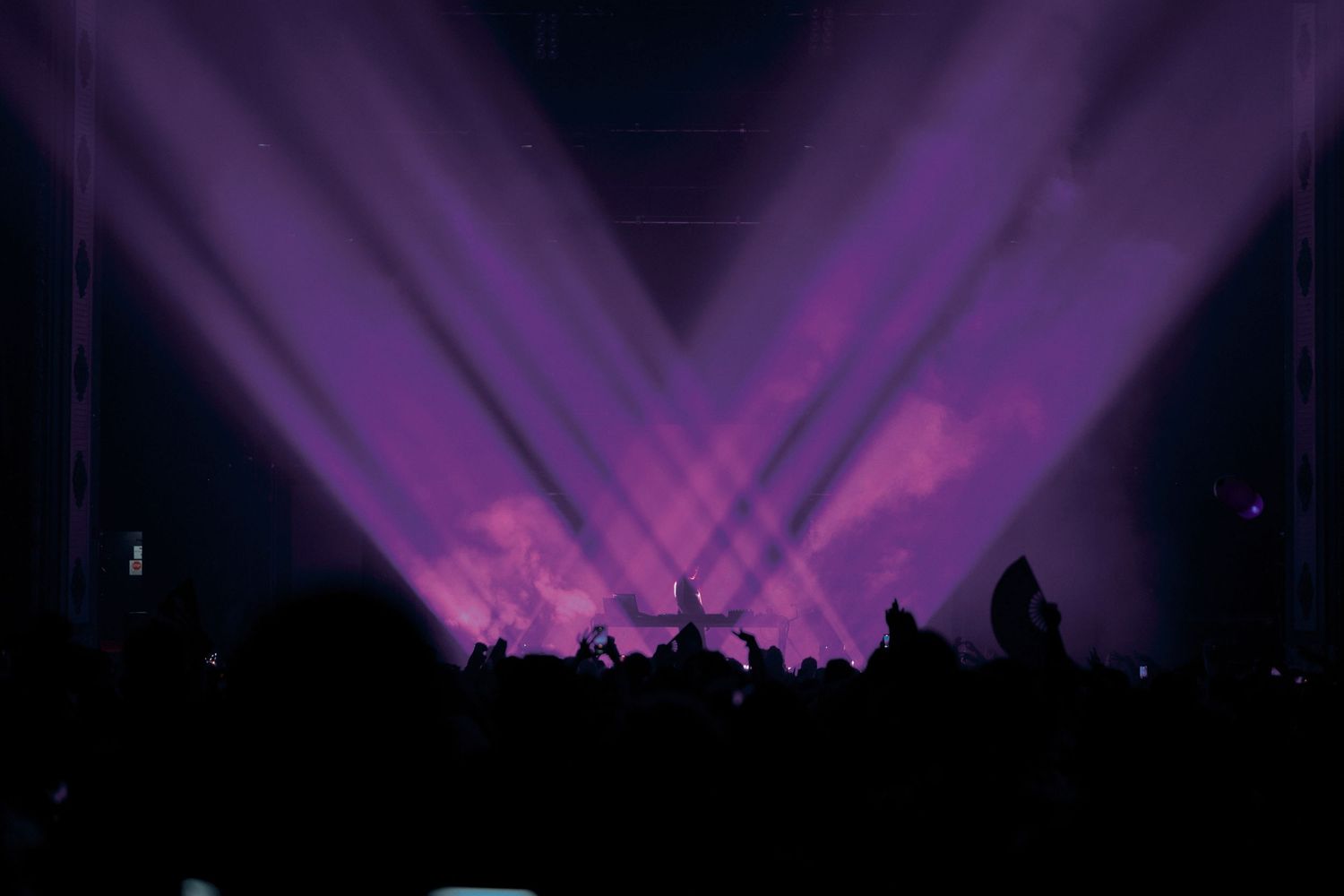 Concert crowd silhouettes against purple stage lights creating dramatic beams in darkness.